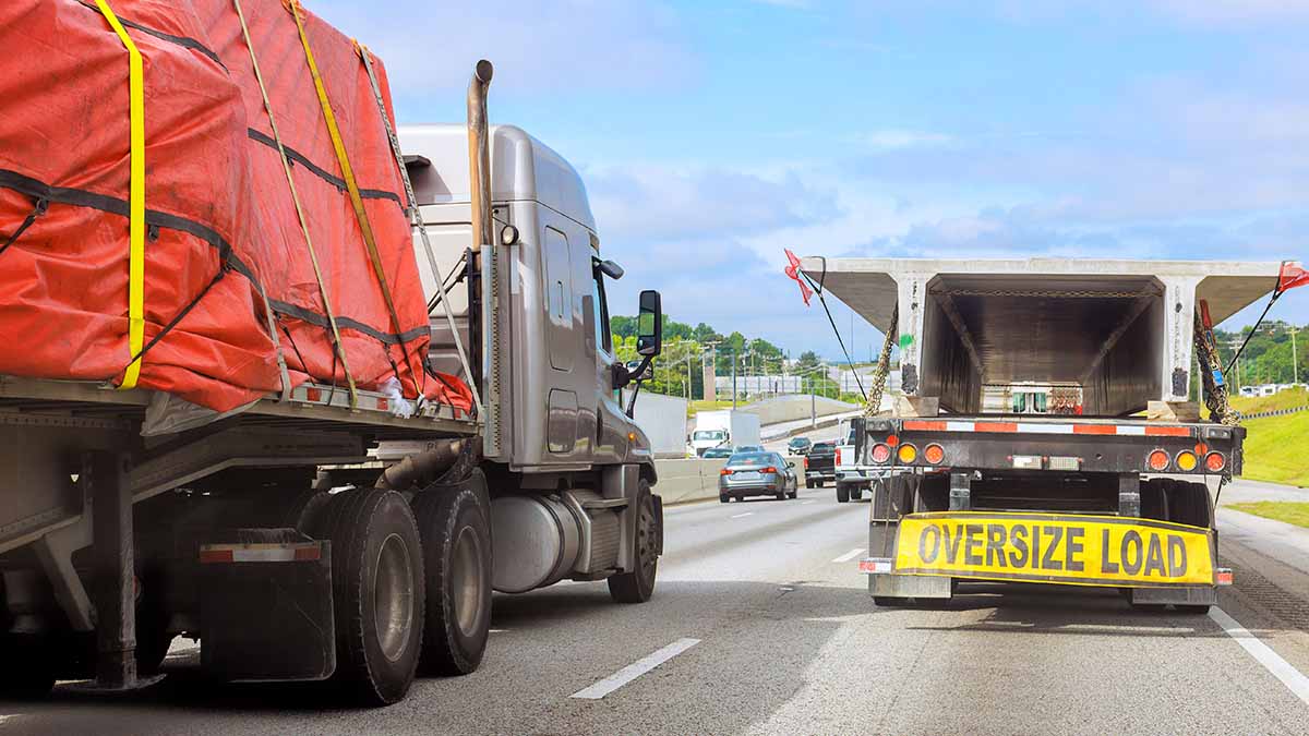 Tractor hauling a tarped oversize load on the interstate, beside a trailer placarded OVERSIZE LOAD.